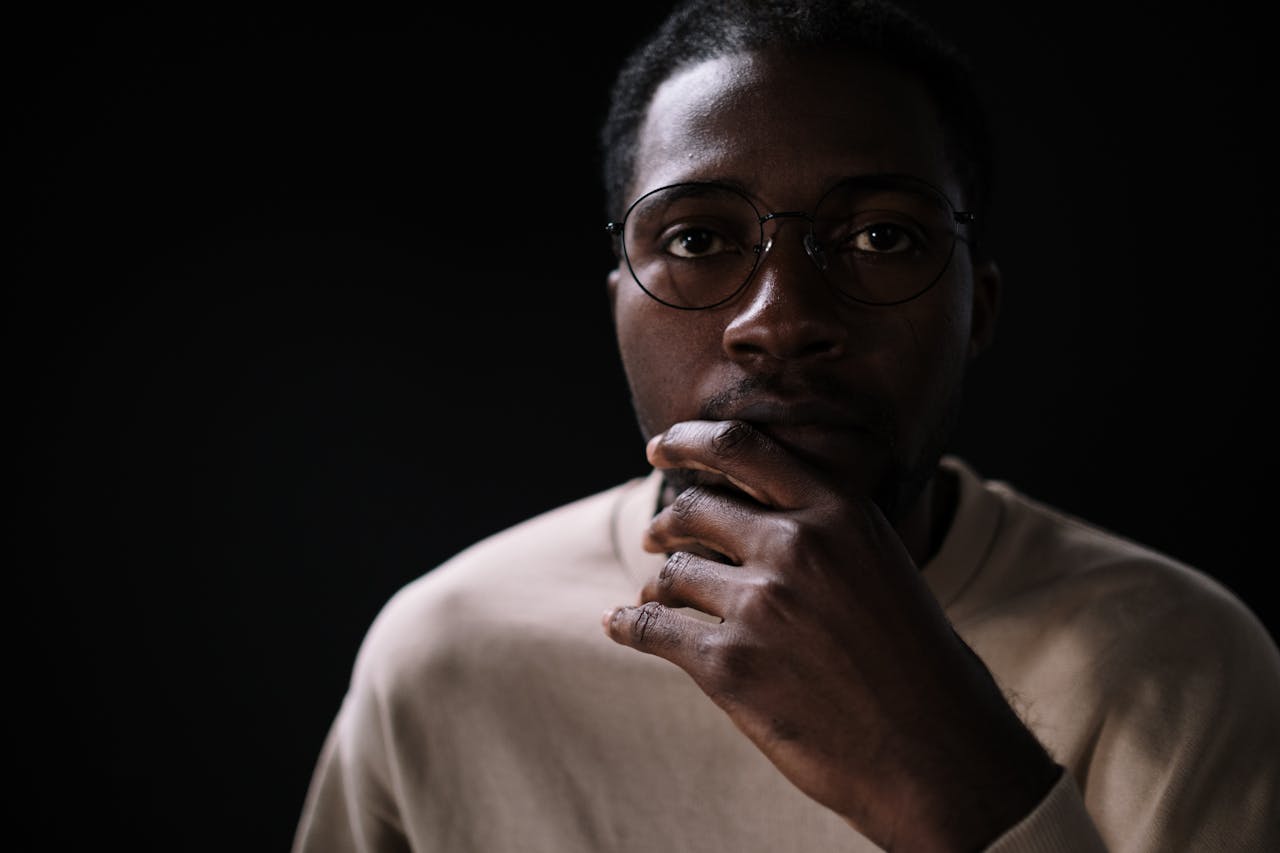 Close-up portrait of a thoughtful African man wearing eyeglasses against a black background indoors.