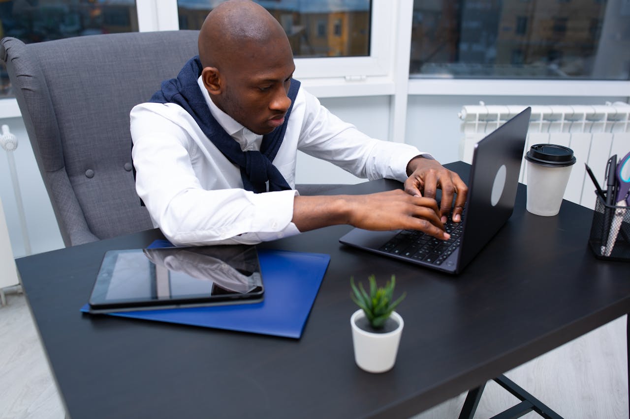 Black man in a white shirt using a laptop at a modern office desk.